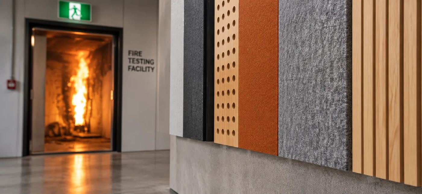 Acoustic panel samples — timber slat, perforated ply, felt and rust-coloured PET — mounted on a wall beside the open door of a fire testing facility with a room test burning inside — Acoustic Panels Australia
